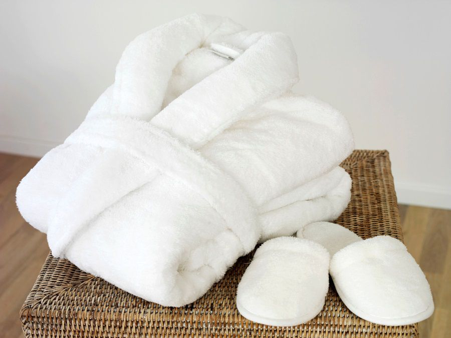 Un peignoir blanc plié très moelleux et des chaussons blancs et doux posés sur un tabouret en osier dans une ambiance spa